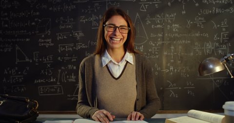 Cinematic shot of young teacher is smiling in camera while correcting exams or homework of students at desk in front of blackboard with math formulas and equations in college or university classroom.