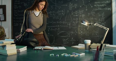 Cinematic shot of young female teacher arrives and sits at her desk to prepare for lecture or lesson in front of blackboard with math formulas and equations in college or university classroom.