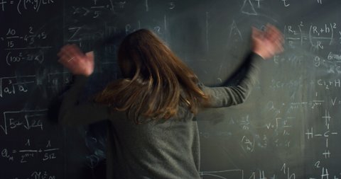 Cinematic shot of young female student writing math formulas and equations on blackboard, cannot find a solution and erases everything with anxiety and frustration in college or university classroom.