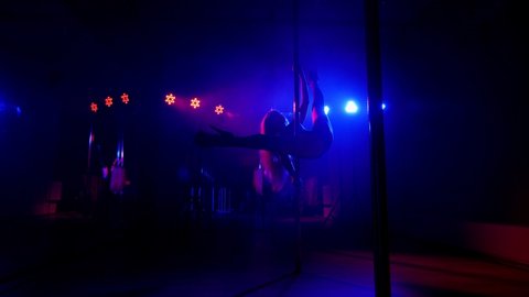 A young, slender woman is dancing on a pole in a dark, smoky room with bright multicolored spotlights. The silhouette of a stripper.