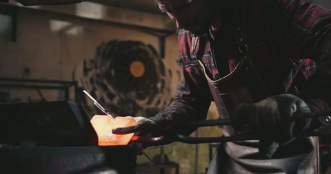 The man holds the hot metal with tongs and shapes it with an automatic machine that hits the workpiece. The blacksmith is wearing protective clothing and earmuffs
