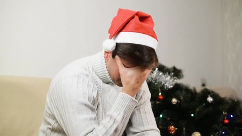 Loneliness in Christmas celebration. Sad, angry, young man in Santa hat in depressed mood sitting alone at home next to fallen decorated Christmas tree and aggressively hitting toys