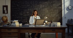 Portrait of Young Female Teacher in Classroom Using Laptop to Give Remote Classes In Front of a Blackboard with Formulas. Science Professor Teaching Online, Having a Video Conference with Students