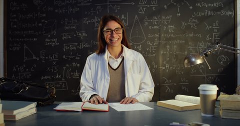 Cinematic shot of young female science professor in lab coat smiling in camera while preparing for lecture at desk in front of blackboard with formulas and equations in college or university classroom