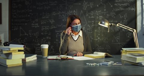 Cinematic shot of young female teacher is taking off protective medical mask and smiling in camera at desk in front of blackboard with math formulas and equations in college or university classroom.