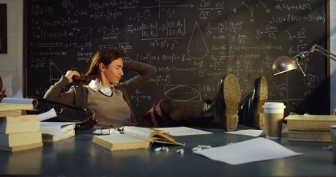 Cinematic shot of young female teacher is relaxing by putting her feet on desk and looking smartphone after finishing lectures in front of blackboard with formulas in college or university classroom.