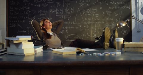 Cinematic shot of young female teacher is relaxing by putting her feet on desk after finishing lectures in front of blackboard with formulas in college or university classroom.