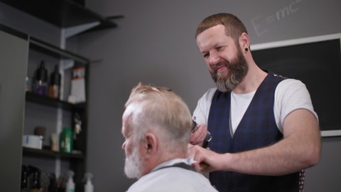 professional male haircutter gives a haircut with a comb and trimmer to a stylish male retiree in a popular barbershop