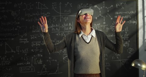 Cinematic shot of female teacher or student watching project by using vr glasses with innovative augmented reality technology in front of blackboard with formulas in college or university classroom.