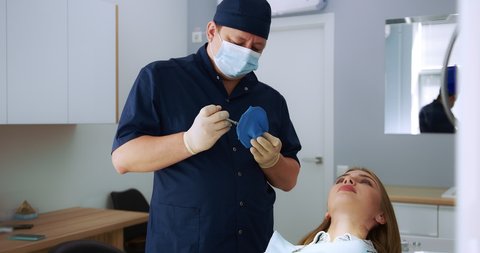 A male dentist in a medical uniform, gloves and mask performs the procedure of applying a cofferdam to a beautiful young woman patient in a modern dental office. High-quality 4k video.