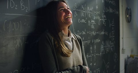 Cinematic close up shot of young happy dreamy female teacher or student leaning against blackboard with formulas and smiling satisfied in college or university classroom with sunshines from window.
