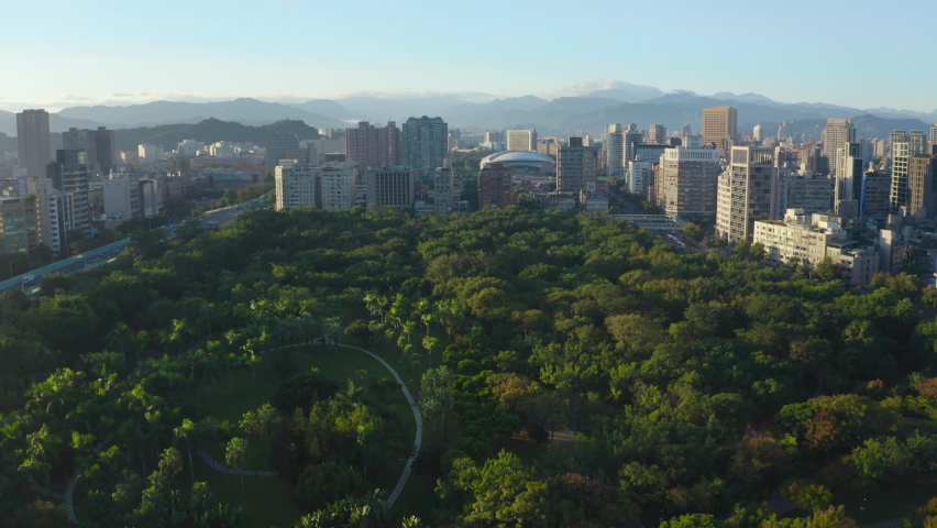 Epic Aerial flyover rural Daan Park with green trees surrounded by skyline with skyscraper buildings and silhouette of mountains in background - Taipei City,Taiwan during daytime and sun