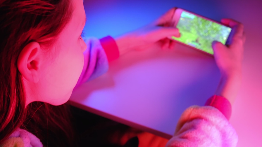 Closeup portrait of pretty young girl playing online mobile game on a smart phone. Teen girl play the game on cellphone. Gamer playing online game on smartphone in dark room. Neon blue and pink light.