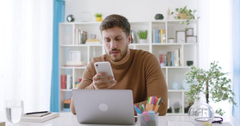 Lateral Dolly of Young Caucasian Bearded Millennial Professional Male Holding Modern Smartphone Texting Message At Home. Thoughtful Cheerful Young Businessman Writing A Text Sitting at Work Desk.