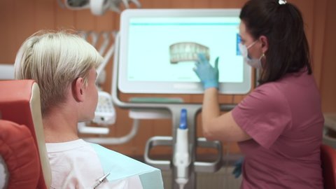 Teenager visits orthodontist. Skilled doctor in pink uniform shows jaws model on computer display to teen blond boy visiting hospital office