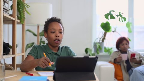 Online education at home. Positive African-American boy draws picture in sketchbook talking to classmates via tablet while mother rests on sofa at home
