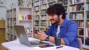 Young Indian male student wearing headset talking and gesturing looking at camera webcam sitting in library making distance video conference call. Online lesson, job interview or vlog recording