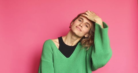 Lovely coquettish brunette woman, winking playfully, flirting, throws her hair. Woman fix her hair at morning. Curly hair fashion model face smiling. Girl in green sweater isolated on pink background.