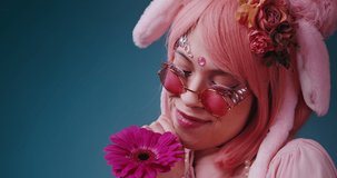 Close up of happy pink cute candy anime girl with rabbit ears and pink sunglasses sniffs a flower and emotional grimaces on blue background. Beautiful funny asian cosplay anime teen girl concept