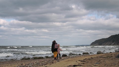 Mother and daughter woman in warm outfit with backpack walk on empty beach at overcast windy morning