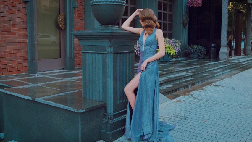 Happy beautiful fashion model woman posing standing on backdrop modern city building. Gray blue evening elegant shiny sexy dress. Retro hairstyle, red long wavy hair. Delicate makeup smiling cute face