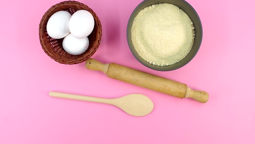 hand puts kitchen tools white eggs and dark gray bowl with whole wheat flour on a pink background
