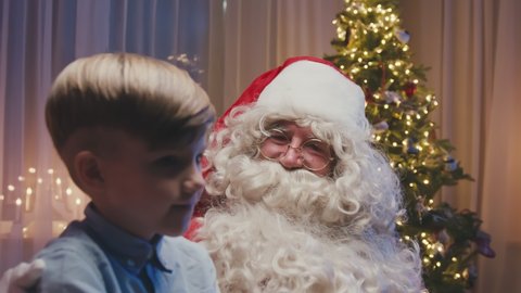 Close up of little cute boy sitting on Santa's knees making a wish. Whitebearded Santa is hugging him and asking him questions.