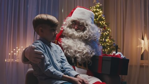 Little cute boy sitting near fireplace on Santa's knees, making a wish. Whitebearded Santa is hugging him and holding a gift.