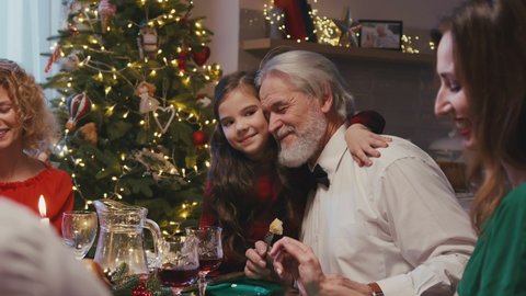 Happy caucasian grandfather sitting at the dinner table with his family celebrating Christmas. Little shy granddaughter giving huggs to grangfather while having Christmas dinner at home.