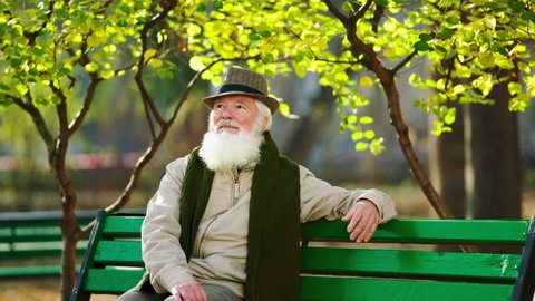 Attractive old man have a walk in the park alone he take a sit down on the chair and enjoy the time in the late autumn day he wearing a cute vintage hat