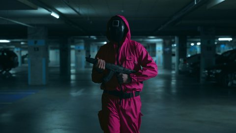 NEW YORK, NY, USA - November 10, 2021: Guard from serial of squid game walking on underground parking with real weapon in hands. Male person wearing red overalls and black mask with white square
