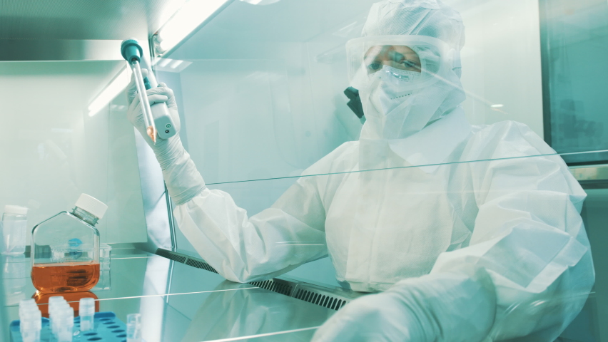 Handheld shot of a Caucasian female lab scientist, in a full protective coverall with a hood, filling test tubes with a pipette in a laminar flow cabinet. Royalty-Free Stock Footage #1082649775