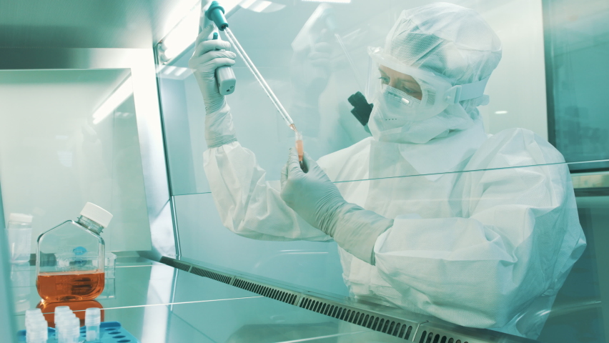 Handheld shot of a Caucasian female lab scientist, in a full protective coverall with a hood, filling test tubes with a pipette in a laminar flow cabinet. | Shutterstock HD Video #1082649775