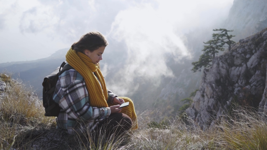 Brunette woman tourist with modern smartphone with long yellow scarf sitting on edge of steep cliff against mountain hidden by deep mist and valley below on sunny autumn day, one with nature