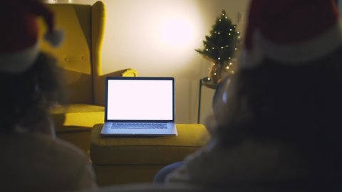 Defocused on foreground couple in Santa hats sitting in front of laptop celebrating happy new year online, cheers with glasses enjoying togetherness at home during coronavirus outbreak