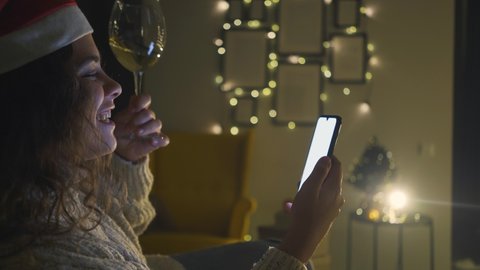 Portrait cheerfull lady in coloured winter festive Santa hat holds champagne wineglass and talks on video call with smartphone in hand against light decorations at home, celebrating online