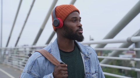 Handsome guy listening to music with headphones and smiling while walking on city bridge. American African person enjoys sounds in earphones and looks at cityscapes with happy smile