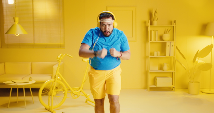 Slow motion of bearded young man doing exercise by running in place and punching movement while wearing headphone to listen to music at home with monochrome yellow interior room background. Royalty-Free Stock Footage #1082650921