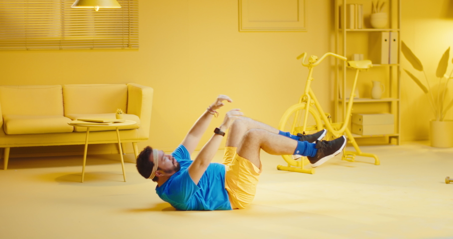 Exhausted overweight bearded young man doing sit ups and taking a break while lying on the floor at home with monochrome yellow interior. Full length shot of a fat male model doing sit up exercise Royalty-Free Stock Footage #1082650960