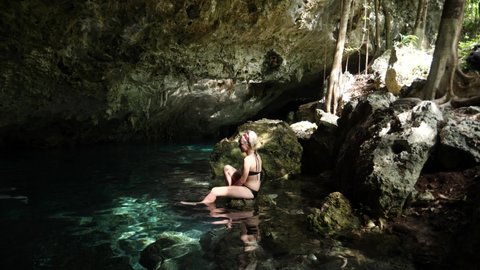 Young blonde woman seating on a rock in a Cenote in Tulum. Turquoise pool in breathtaking underground cave. Natural pool of a sinkhole in Mexico. Beautiful underground Mexican Cenotes.