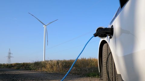 Windmill generates alternative energy for ev in field. Electric ev car with connected plug stands near rotating propeller against high voltage line