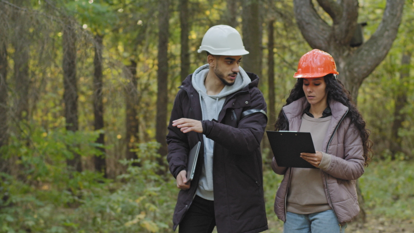 Young diverse employees in helmets, Indian man and oriental woman, forestry engineers in hardhats with tablet walking in park, checking trees talking planning measures for reforestation of woodlands Royalty-Free Stock Footage #1082658445