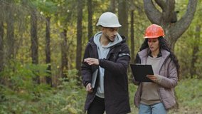 Young diverse employees in helmets, Indian man and oriental woman, forestry engineers in hardhats with tablet walking in park, checking trees talking planning measures for reforestation of woodlands - Powered by Shutterstock - Get 15% off with code: PIKWIZARD15