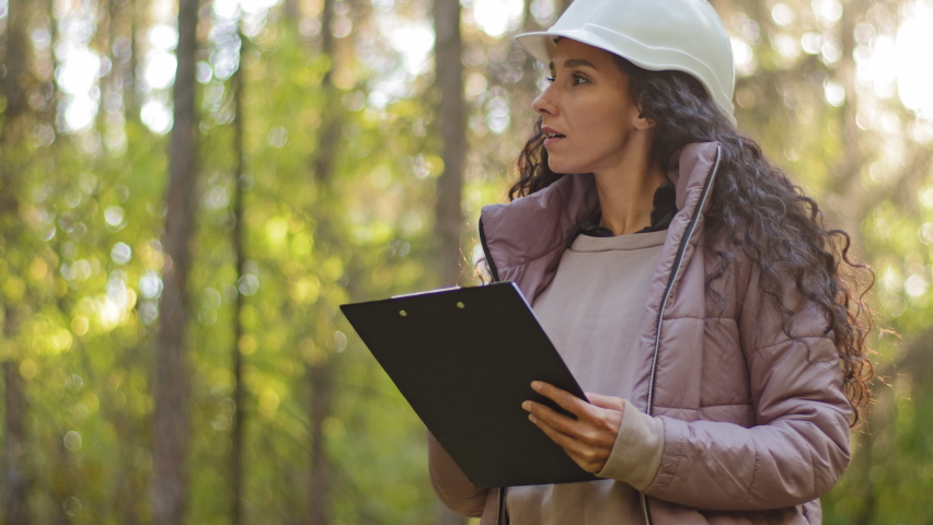 Millennial female technician ecologist looking up at treetops, Young indian woman in hardhat with clipboard taking measures checking trees. Forestry engineer in park. Supervising wildlife sanctuary Royalty-Free Stock Footage #1082659276
