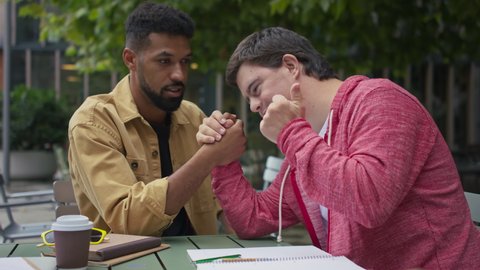 Young man with Down syndrome with his mentoring friend arm wrestling outdoors in cafe
