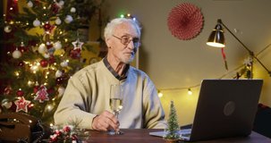 Old man pensioner speaking on video call using laptop, holding champagne. Grandfather talking online and smiling, sitting in decorated living room. Christmas holidays during coronavirus lockdown.