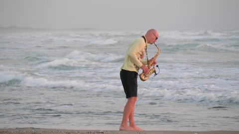 Video In summer, young bald man saxophonist plays golden alt saxophone with microphone, on seashore, beautiful waves with white foam, against sea, in yellow sweater, black shorts. Island of Cyprus.