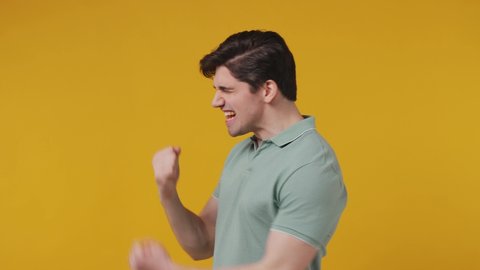 Excited jubilant overjoyed happy joyful attractive happy young man 20s years old wears blue t-shirt doing winner gesture celebrate clenching fists say yes isolated on plain yellow background studio