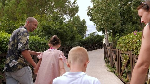 A happy family with children with bare feet are running forward on the bridge among beautiful green bushes and trees. A big happy family on summer vacation.