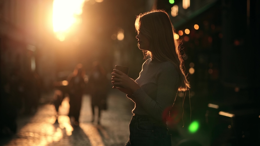 Silhouette of Happy young hipster woman drinking coffee at sunrise. Girl taking a sip of coffee or tea from wooden eco cup on warm summer evening sunset in big city. Natural beauty pretty woman